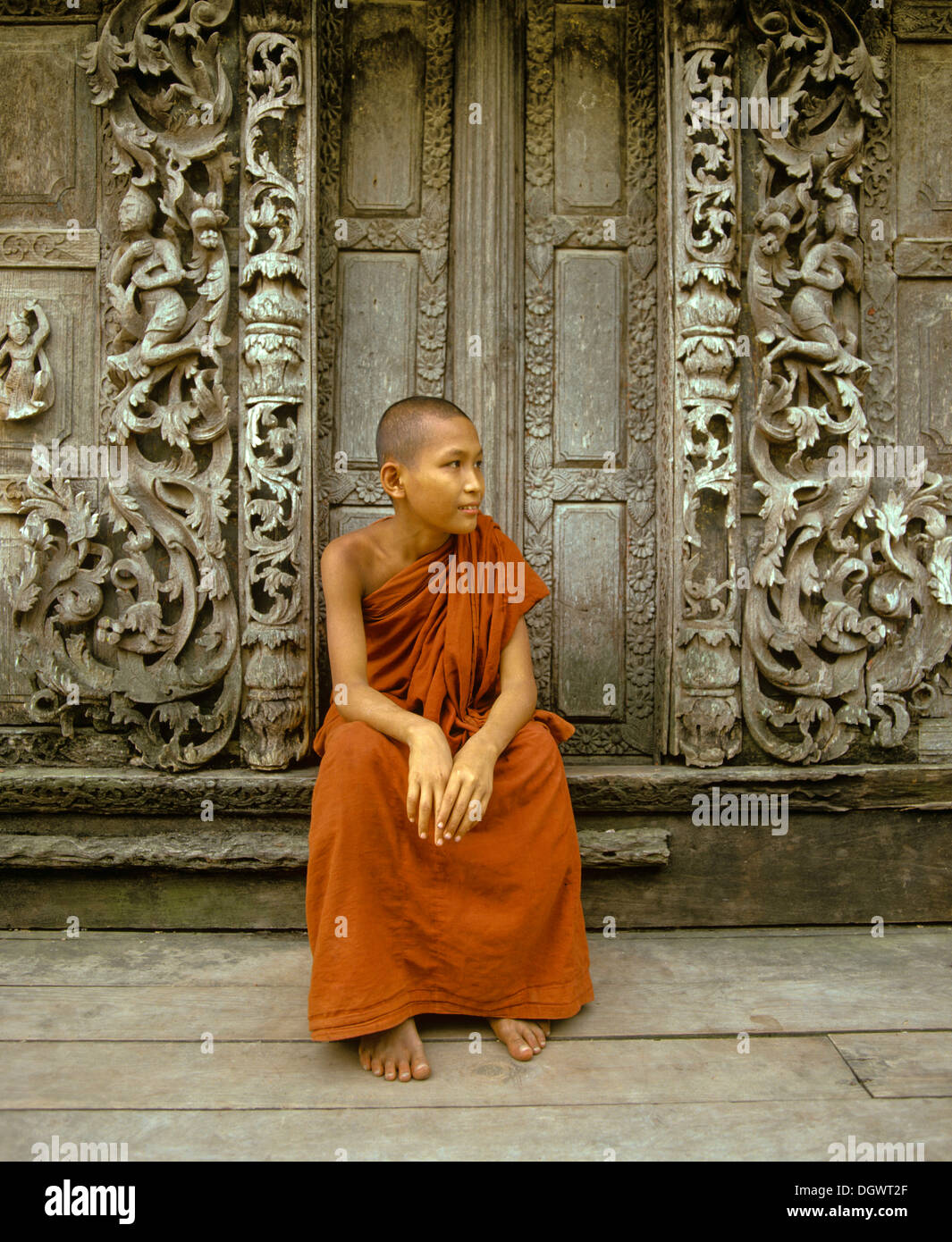 Un moine assis en face d'une porte en bois avec des sculptures en teck, Shwe In Bin Kyaung Monastère, Mandalay Division, Myanmar, Mandalay Banque D'Images