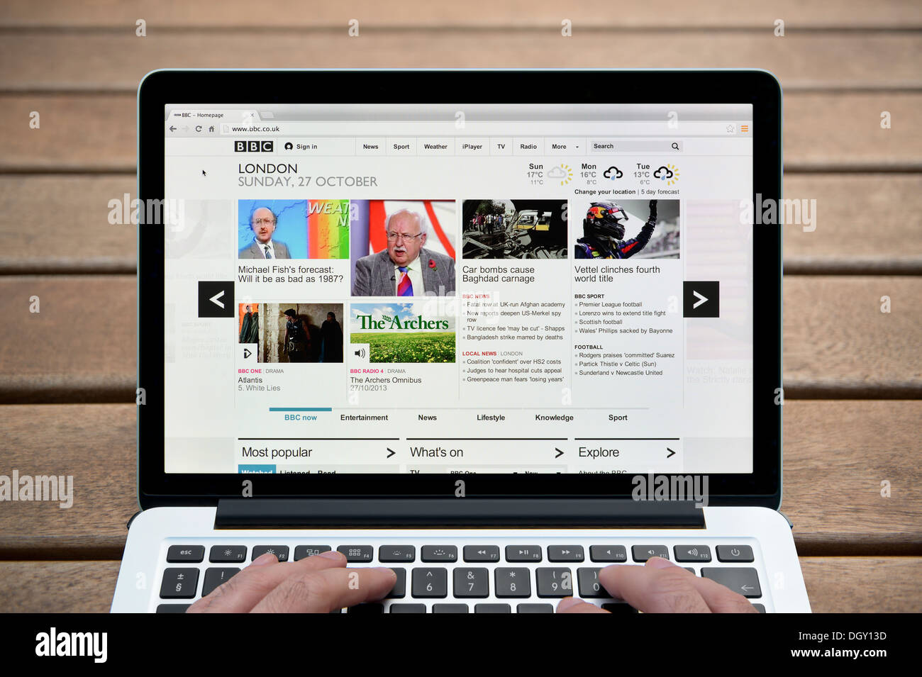 La BBC Online site sur un MacBook contre un banc en bois fond de plein air y compris les doigts d'un homme (usage éditorial uniquement). Banque D'Images