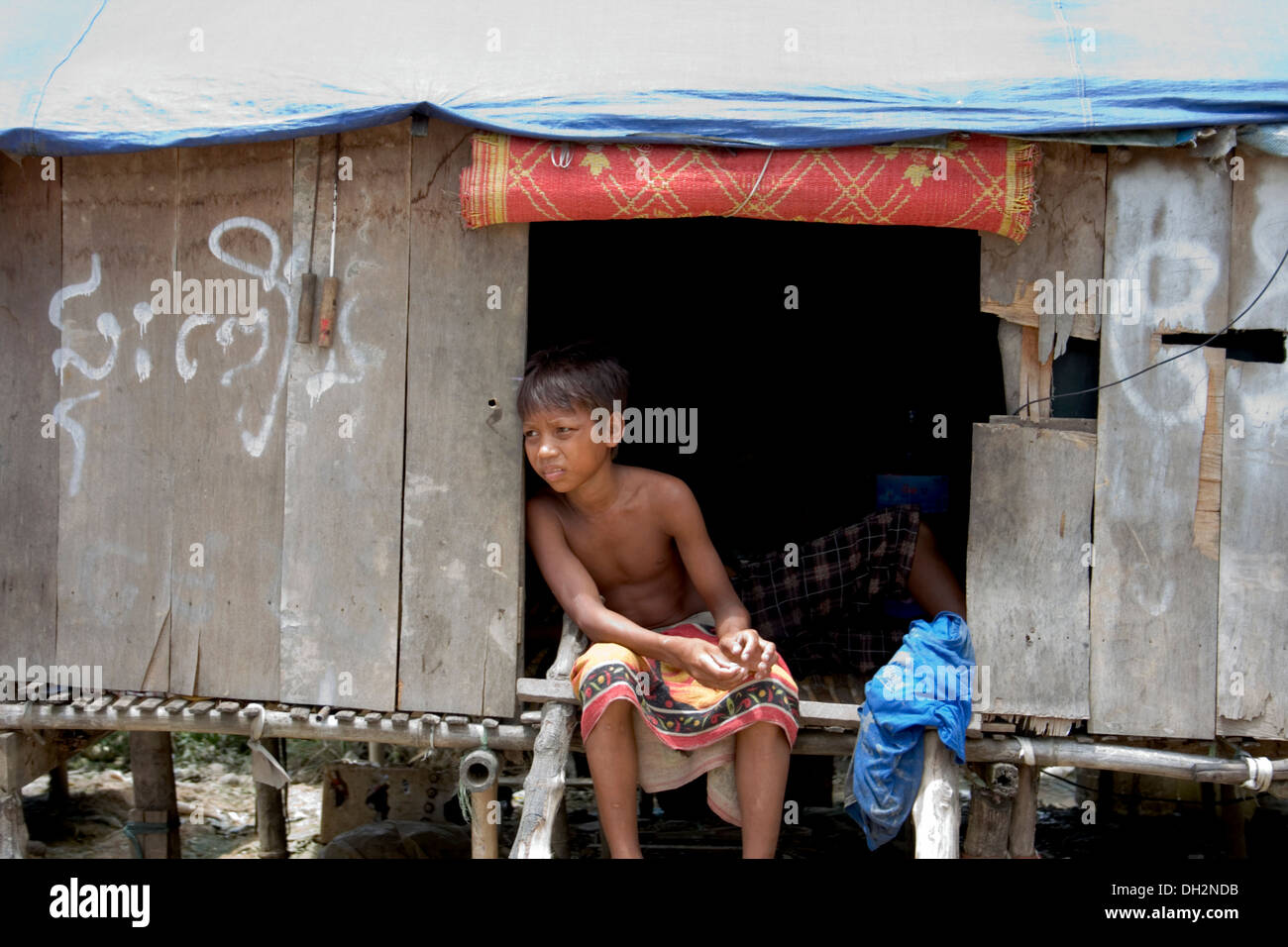 Un enfant travailleur garçon qui travaille dans l'industrie du recyclage est assis sur le seuil de sa maison en bois à Phnom Penh, Cambodge. Banque D'Images
