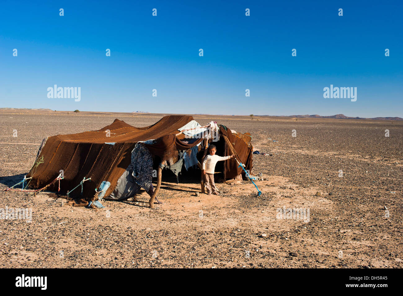 Jeune garçon debout devant la tente de sa famille nomade, pierre désert, Hamada, l'Erg Chebbi, dans le sud du Maroc, Maroc Banque D'Images
