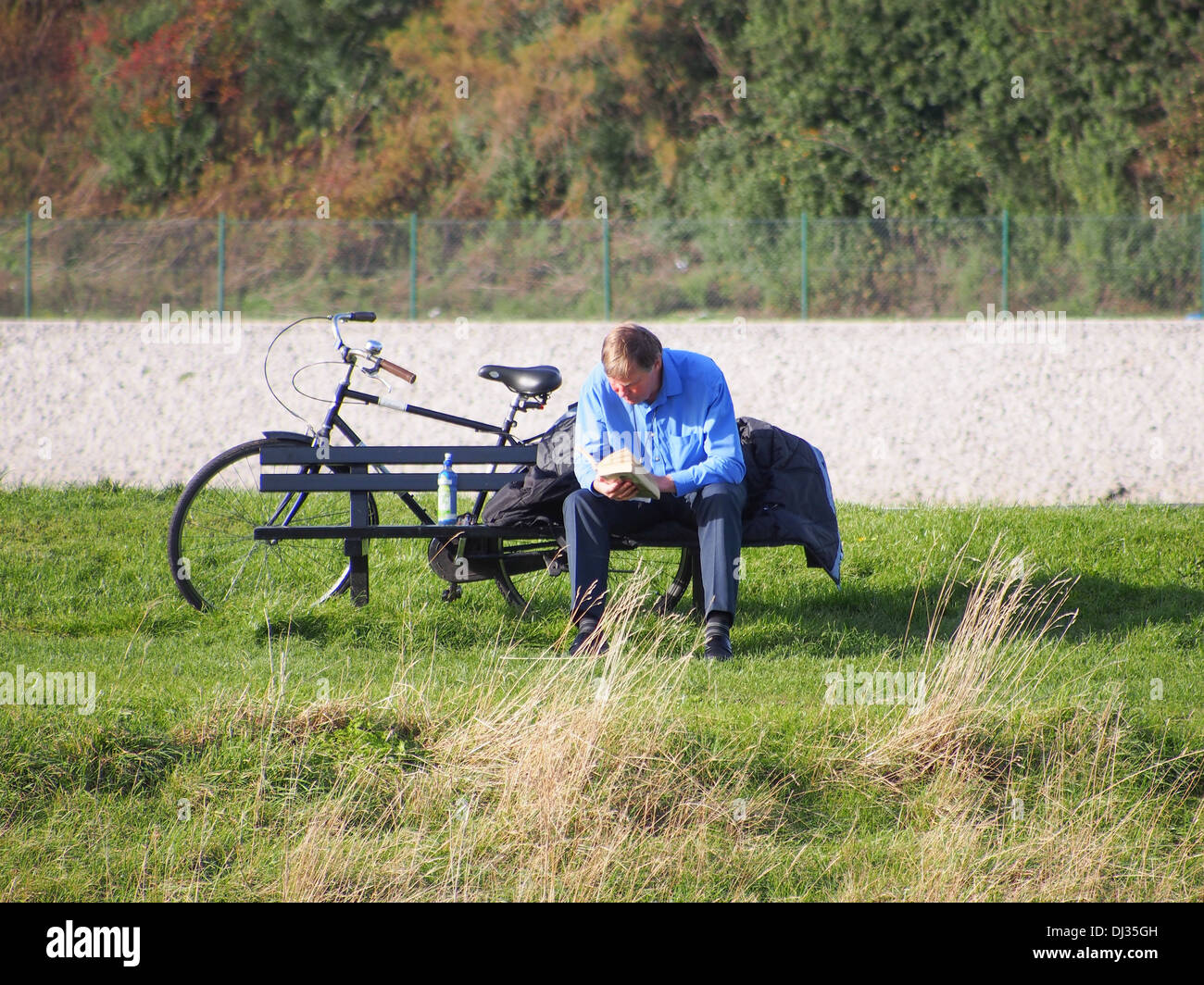 Homme assis sur un banc de parc pour lire un livre avec son vélo derrière lui Banque D'Images