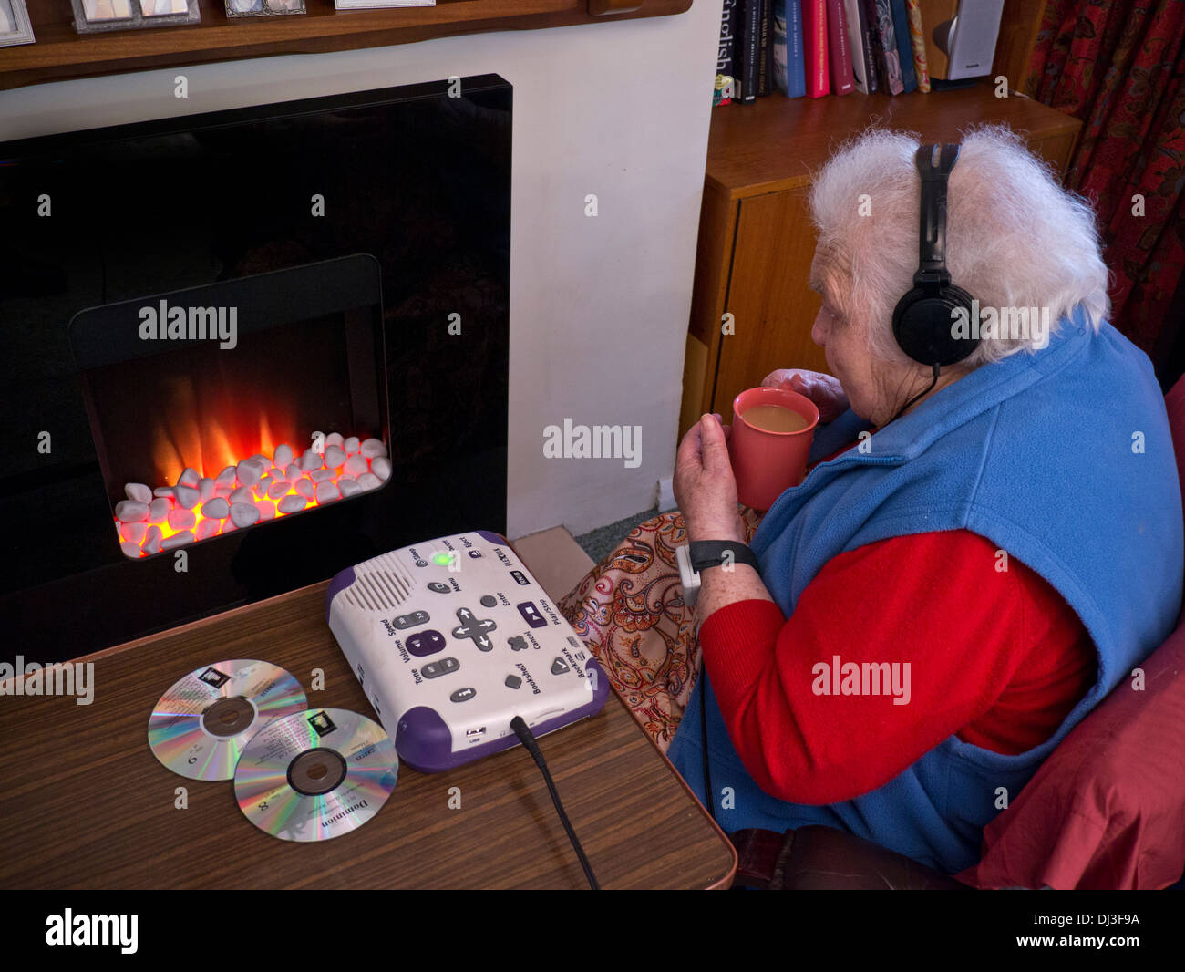 Vieille dame âgée assise isolant covid-19 CORONAVIRUS par un feu de gaz à charbon écoute de livre audio, pour les malentendants, les personnes malvoyantes Banque D'Images