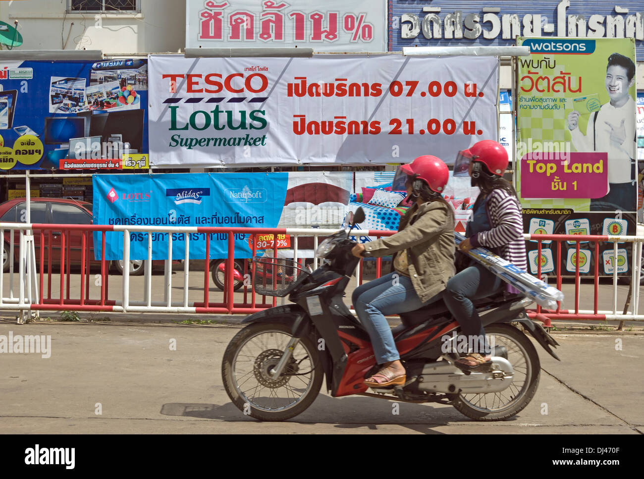 Deux femmes sur une moto passer un signe pour un supermarché Tesco Lotus, avec temps d'ouverture en thaï, à phetchabun, Thaïlande Banque D'Images