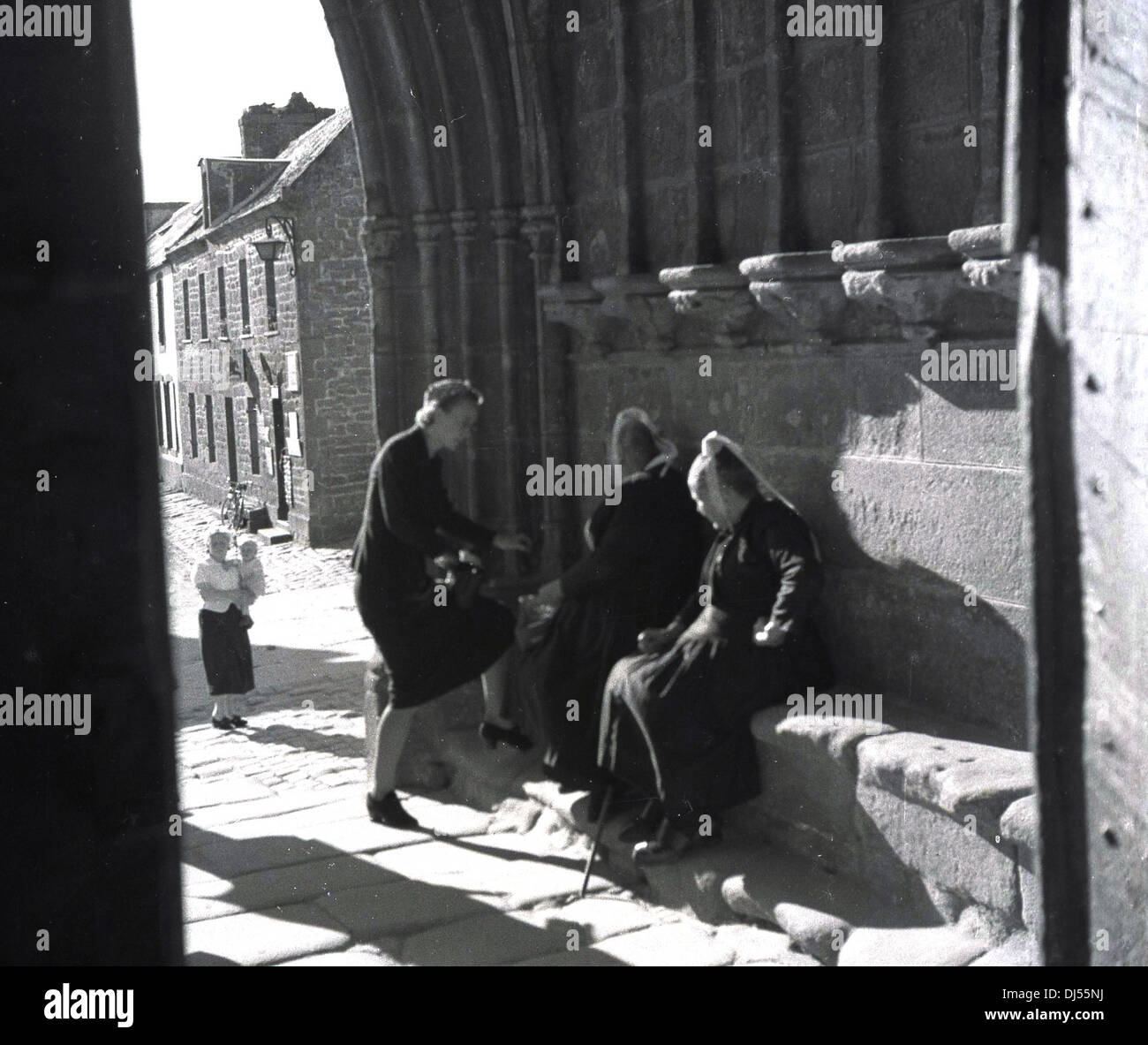 Photo historique de 1930s. Deux religieuses belges assises sur un siège en pierre à l'entrée voûtée d'une église à Liège, Belgique parlant à une dame. Banque D'Images
