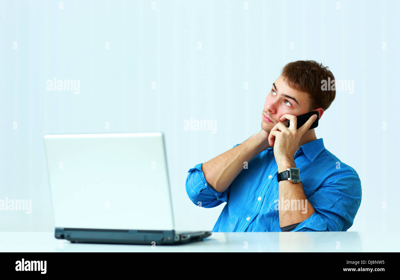 Jeune homme assis à la table avec ordinateur portable et parler au téléphone en fonction Banque D'Images