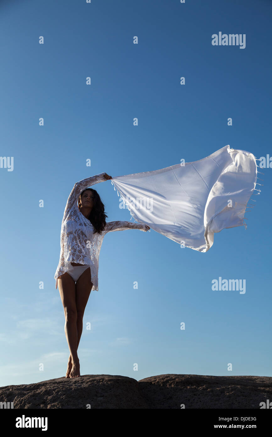 Modèle féminin en agitant une longue écharpe blanche dans l'air contre un ciel bleu profond tandis que se tenait pieds nus sur un rocher wearing white bikini Banque D'Images