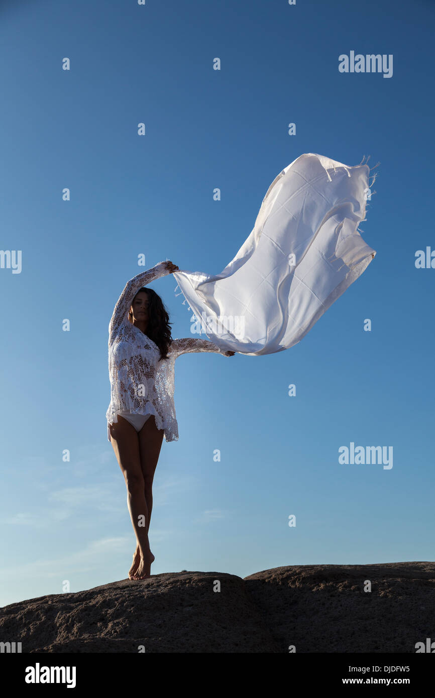 Modèle féminin en agitant une longue écharpe blanche dans l'air contre un ciel bleu profond tandis que se tenait pieds nus sur un rocher wearing white bikini Banque D'Images
