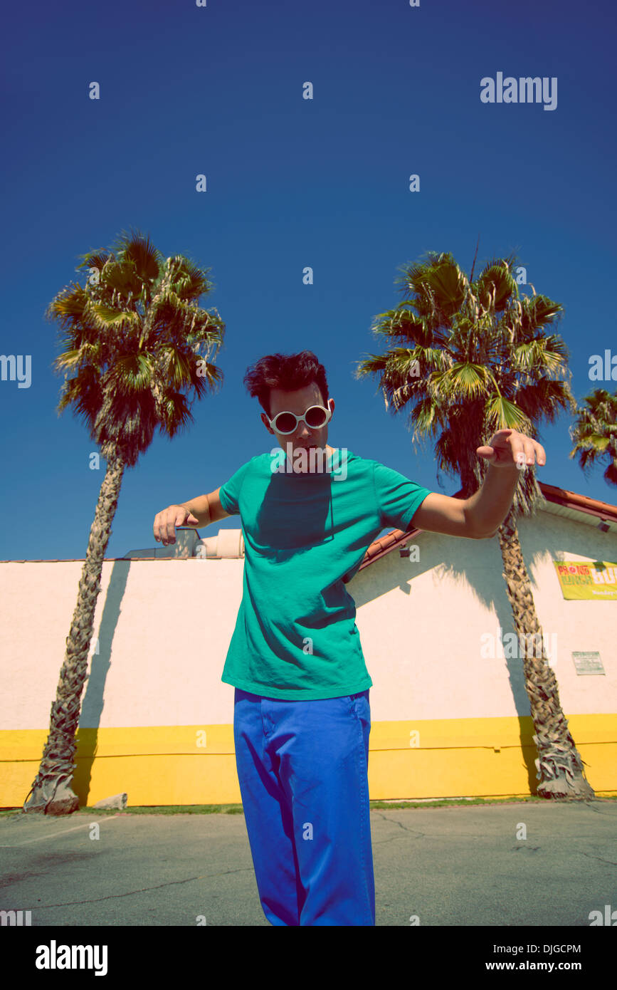 Un jeune homme avec des lunettes rondes et des cheveux funky, permanent, sous ciel bleu entre les palmiers à Los Angeles, Californie Banque D'Images