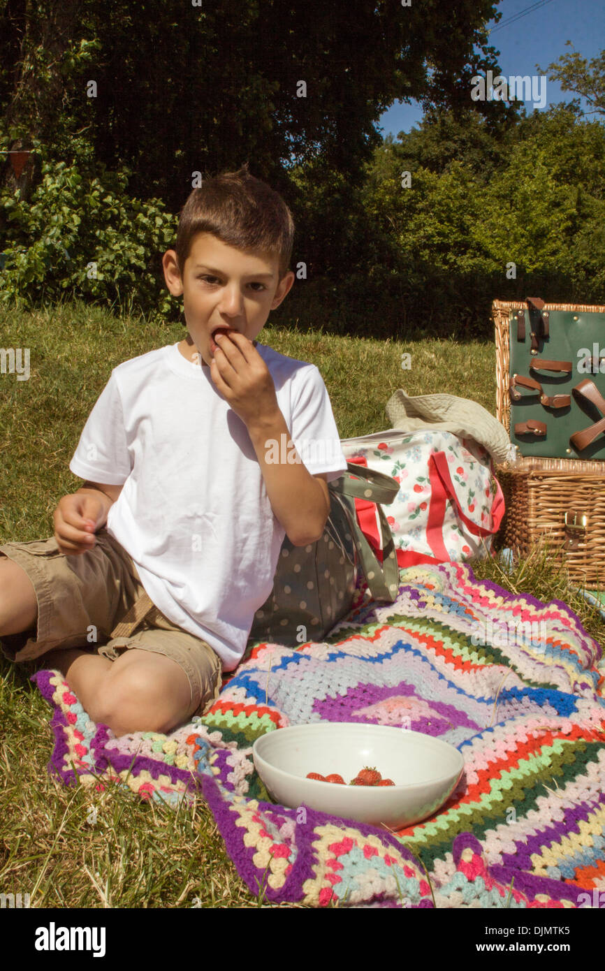 Un garçon sur un pique-nique assis dans des couvertures sur l'herbe de manger des fraises dans le Somerset, Royaume-Uni. Banque D'Images