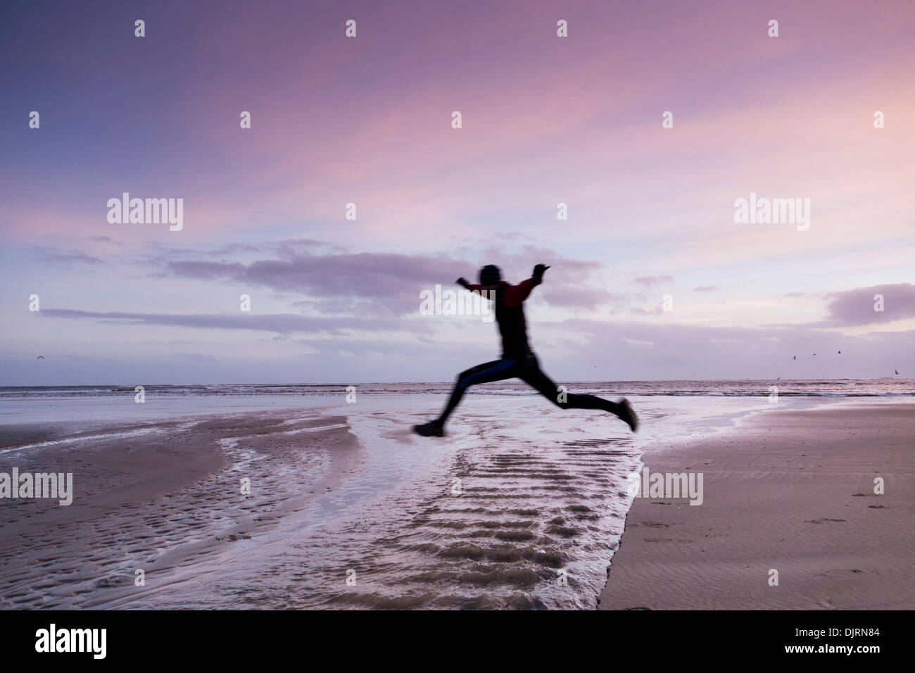 Jogger dans les airs comme il saute au-dessus de l'eau sur la plage au lever du soleil. Angleterre, Royaume-Uni Banque D'Images