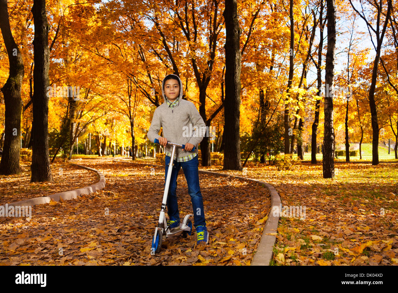 Un beau black 10 ans boy riding a scooter in autumn park avec maple feuilles automne orange portant des sweat avec capuche Banque D'Images