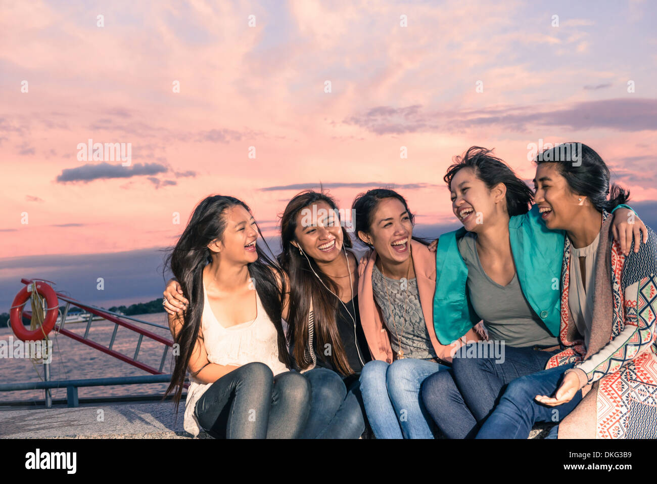 Les jeunes femmes se trouvant assis à bord de l'eau au crépuscule avec les bras autour de Banque D'Images