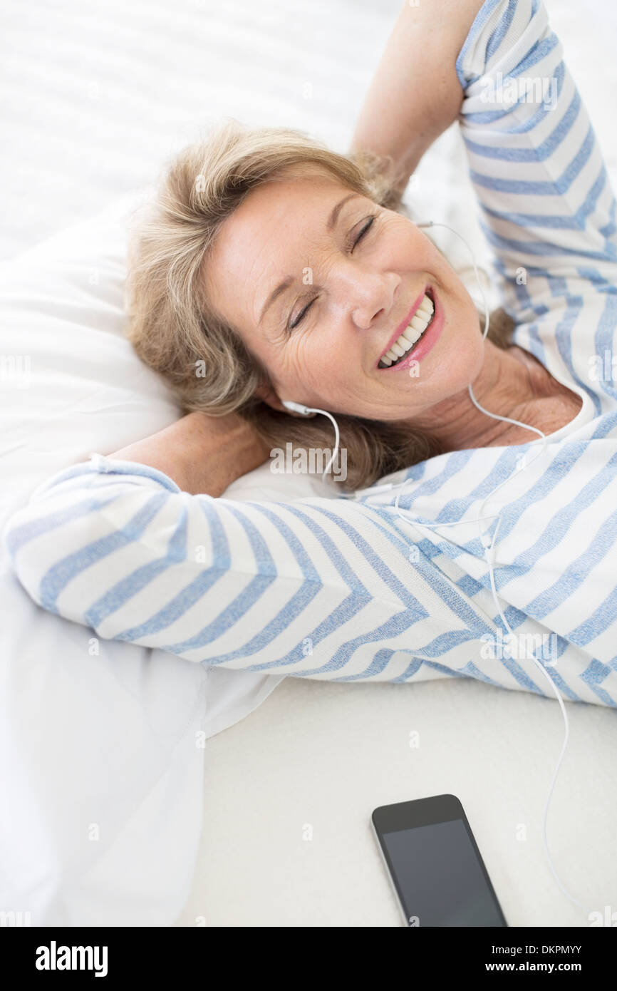Femme plus âgée listening to earphones on bed Banque D'Images