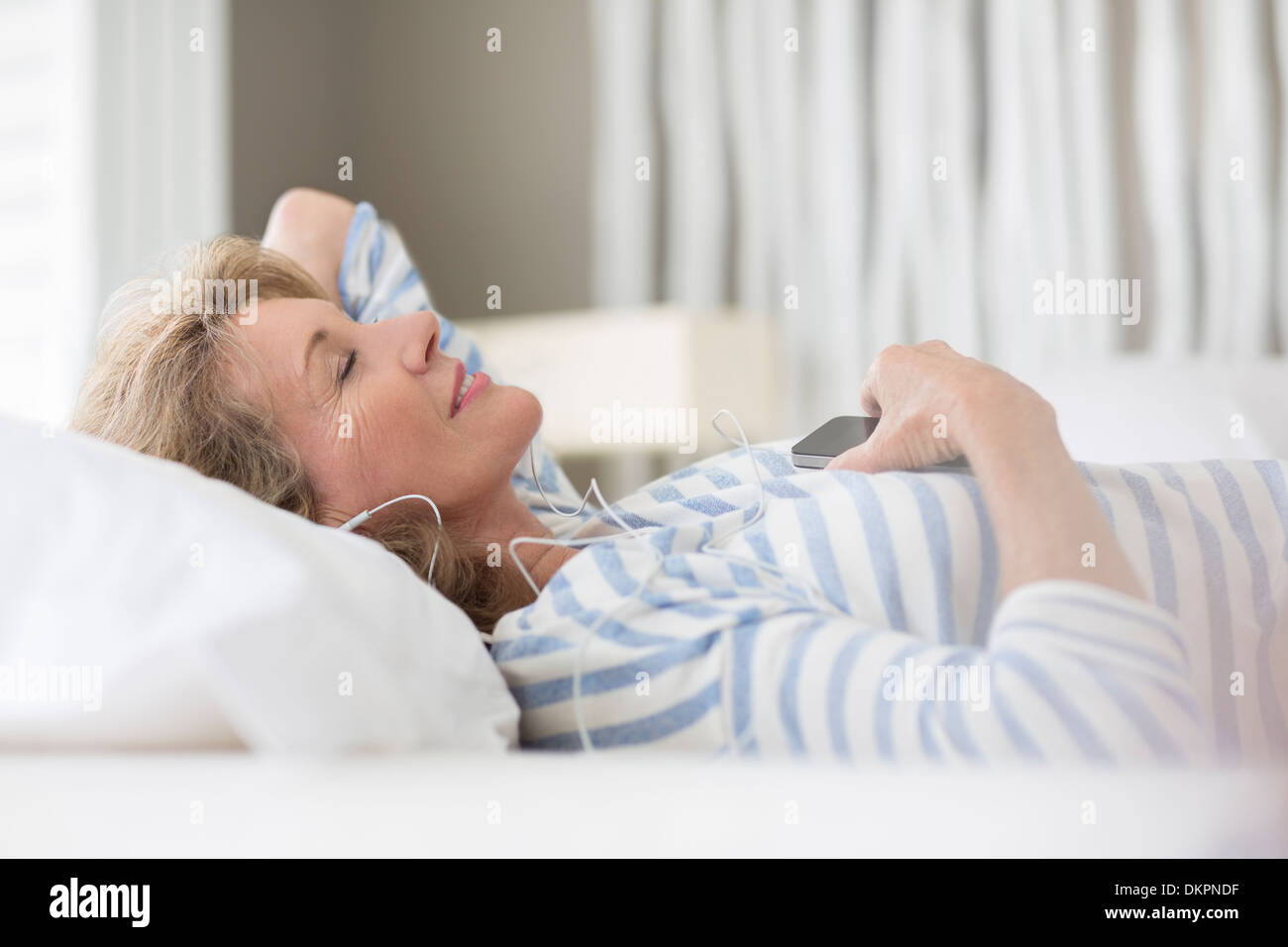 Femme plus âgée listening to earphones on bed Banque D'Images