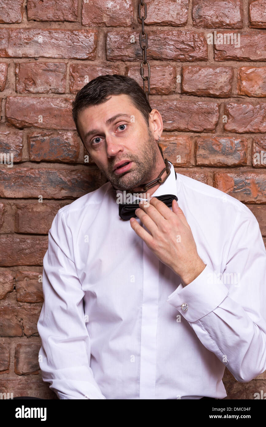 Handsome businessman désespéré dans un nœud papillon debout contre un mur avec une chaîne autour du cou Banque D'Images