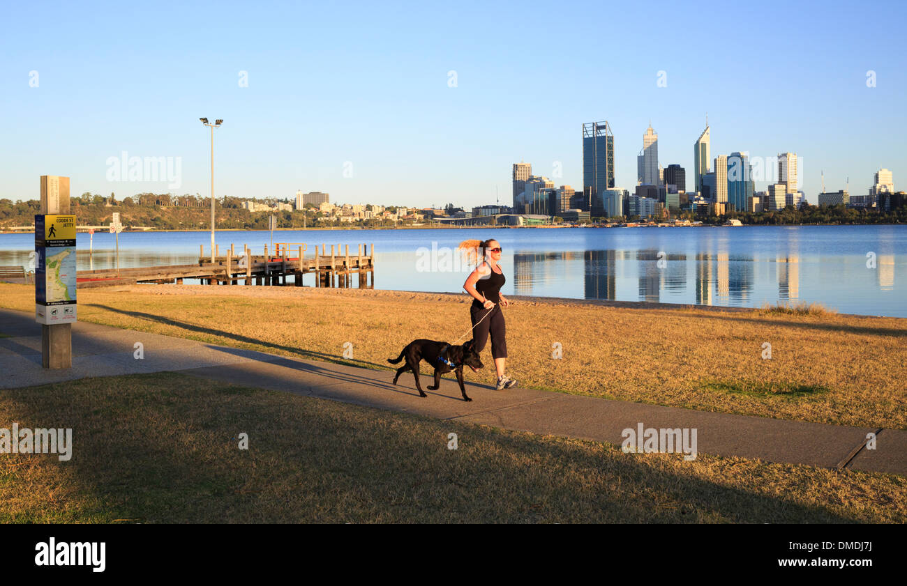 Une femme courir avec un chien le long d'un chemin au bord de la ville avec des gratte-ciel dans le lointain. Perth, Australie occidentale Banque D'Images