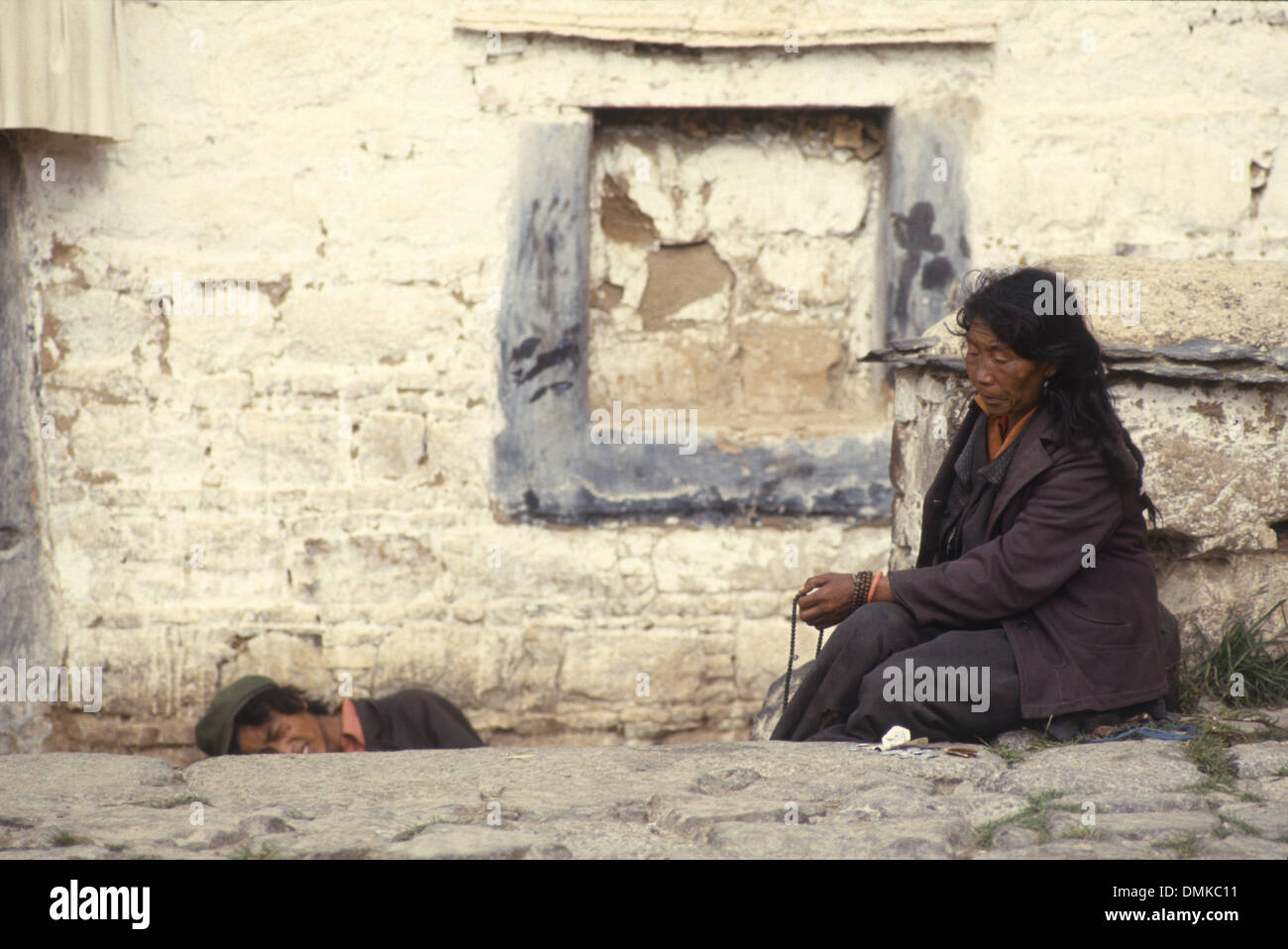 Une pauvre vieille femme assise dans une rue de Lhassa, Tibet, Chine. Un jeune homme se cache de l'appareil photo. Banque D'Images