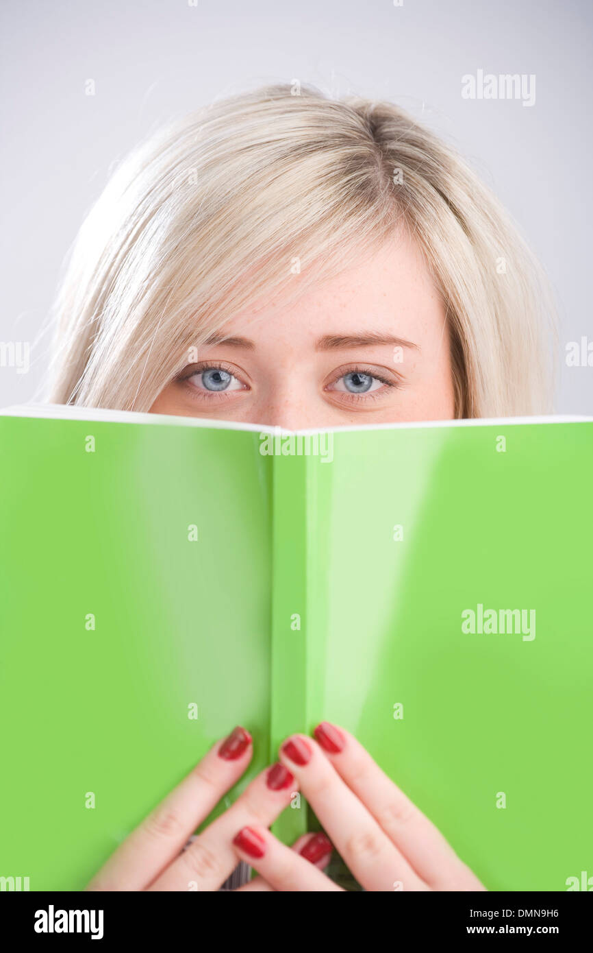 Jolie blonde adolescente peeking sur un livre vert qui a eu lieu en face de son visage. Banque D'Images
