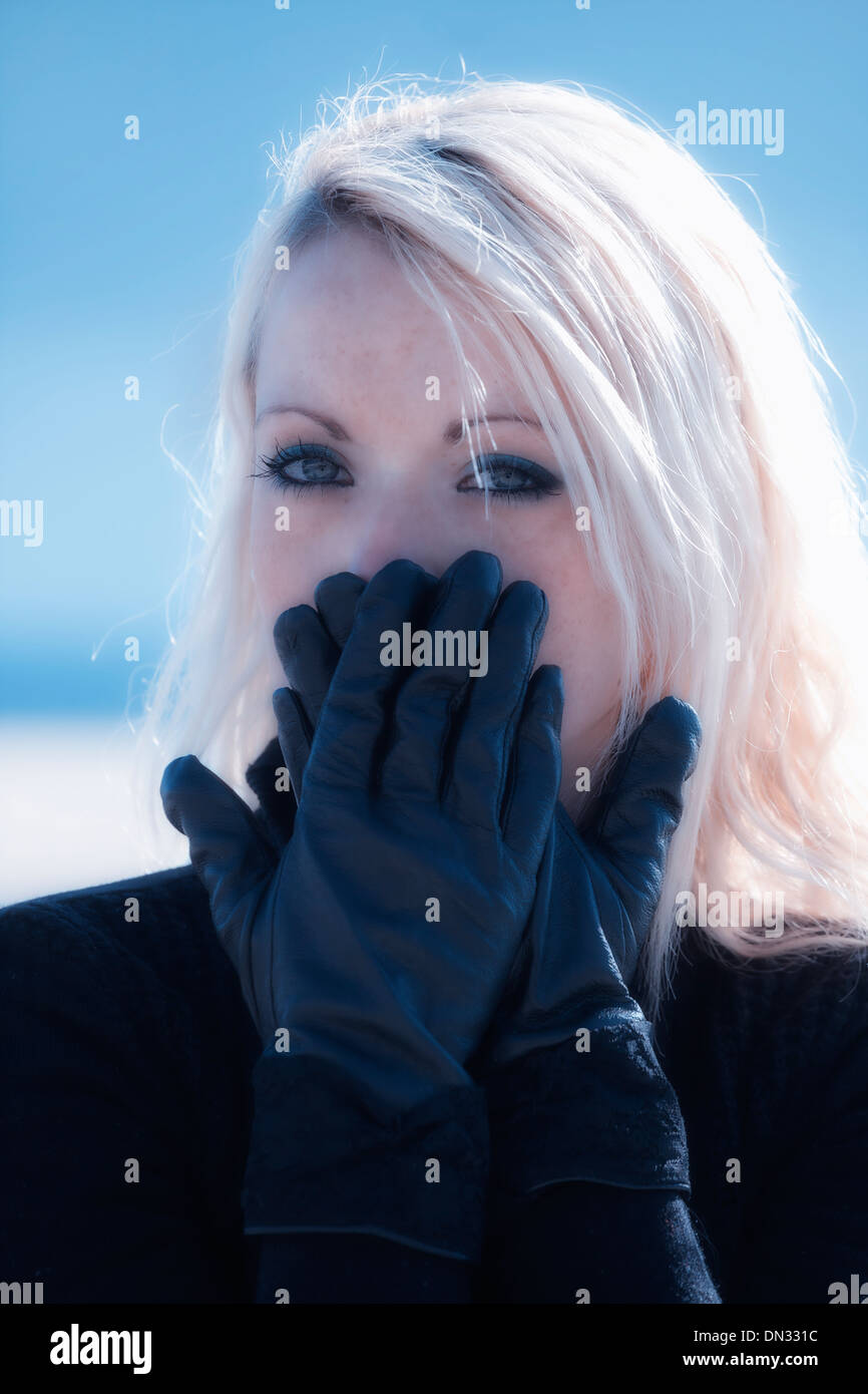 Portrait d'une belle jeune femme avec de longs cheveux blonds qui tient ses mains avec des gants noir devant sa bouche Banque D'Images