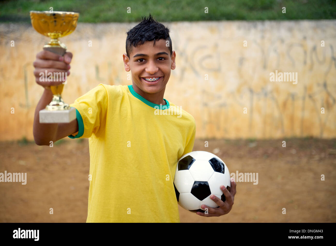Jeune joueur de football brésilien, holding trophy et ballon de soccer sur un terrain de football de saleté de base Banque D'Images