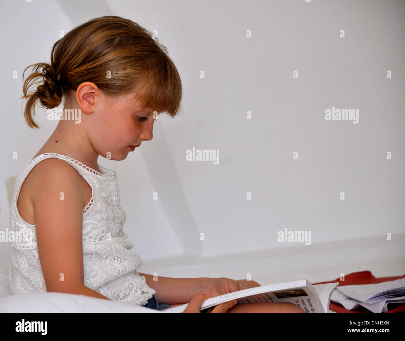 Portrait de profil d'une petite fille blonde vêtue de blanc de la lecture d'un livre sur un fond blanc en vacances Banque D'Images