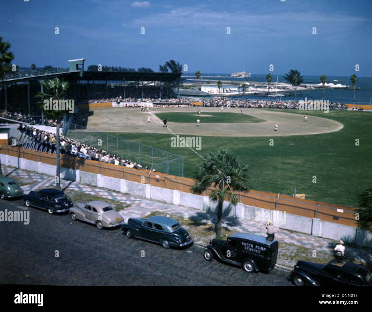 Un match de baseball à Al Lang Field à Petersburg, Floride, dans les années 1940 L'image capte l'énergie du jeu, avec des spectateurs et des voitures classiques en arrière-plan. Banque D'Images