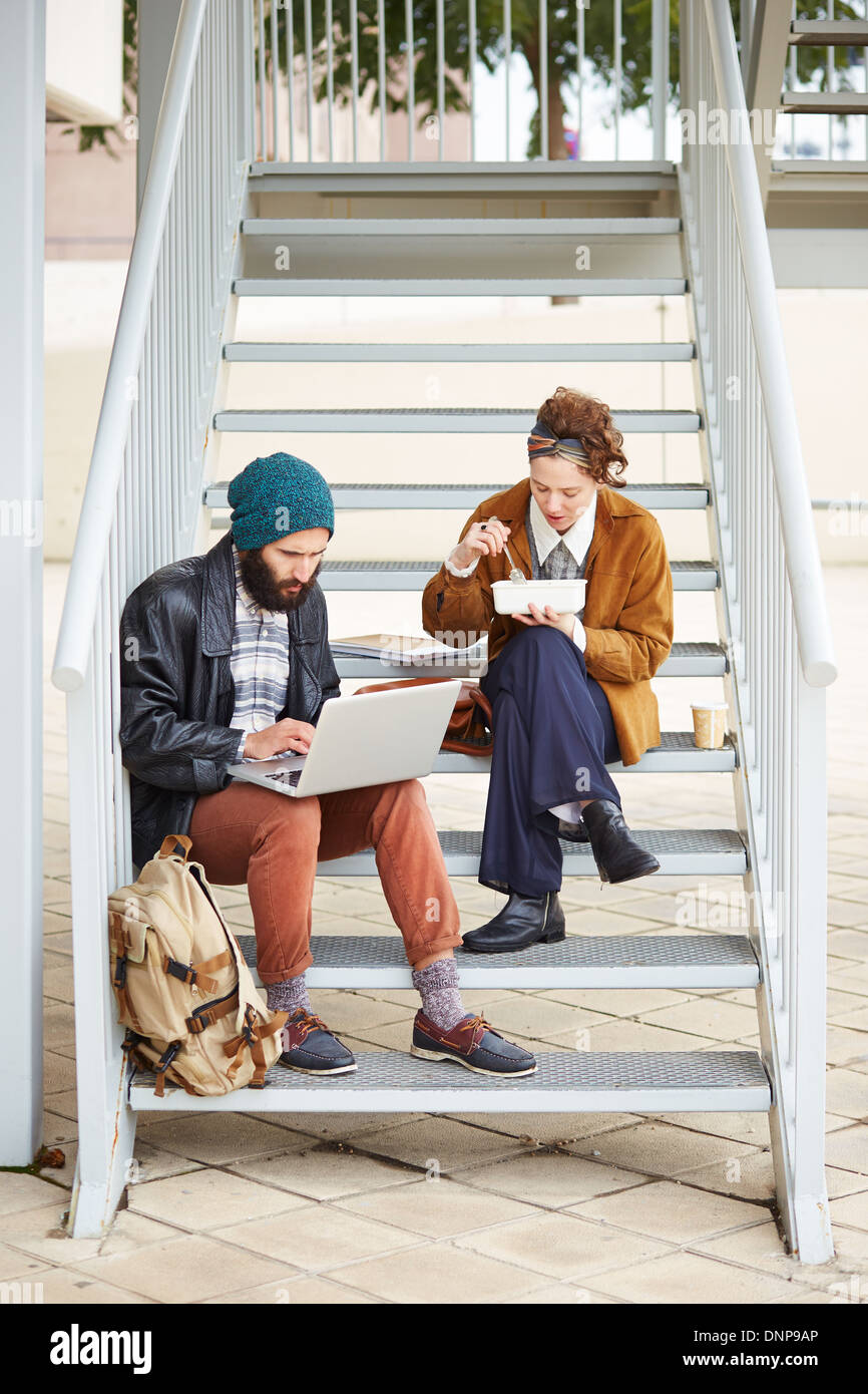 Couple Hipster en utilisant l'ordinateur et manger le déjeuner assis dans les escaliers au campus universitaire Banque D'Images