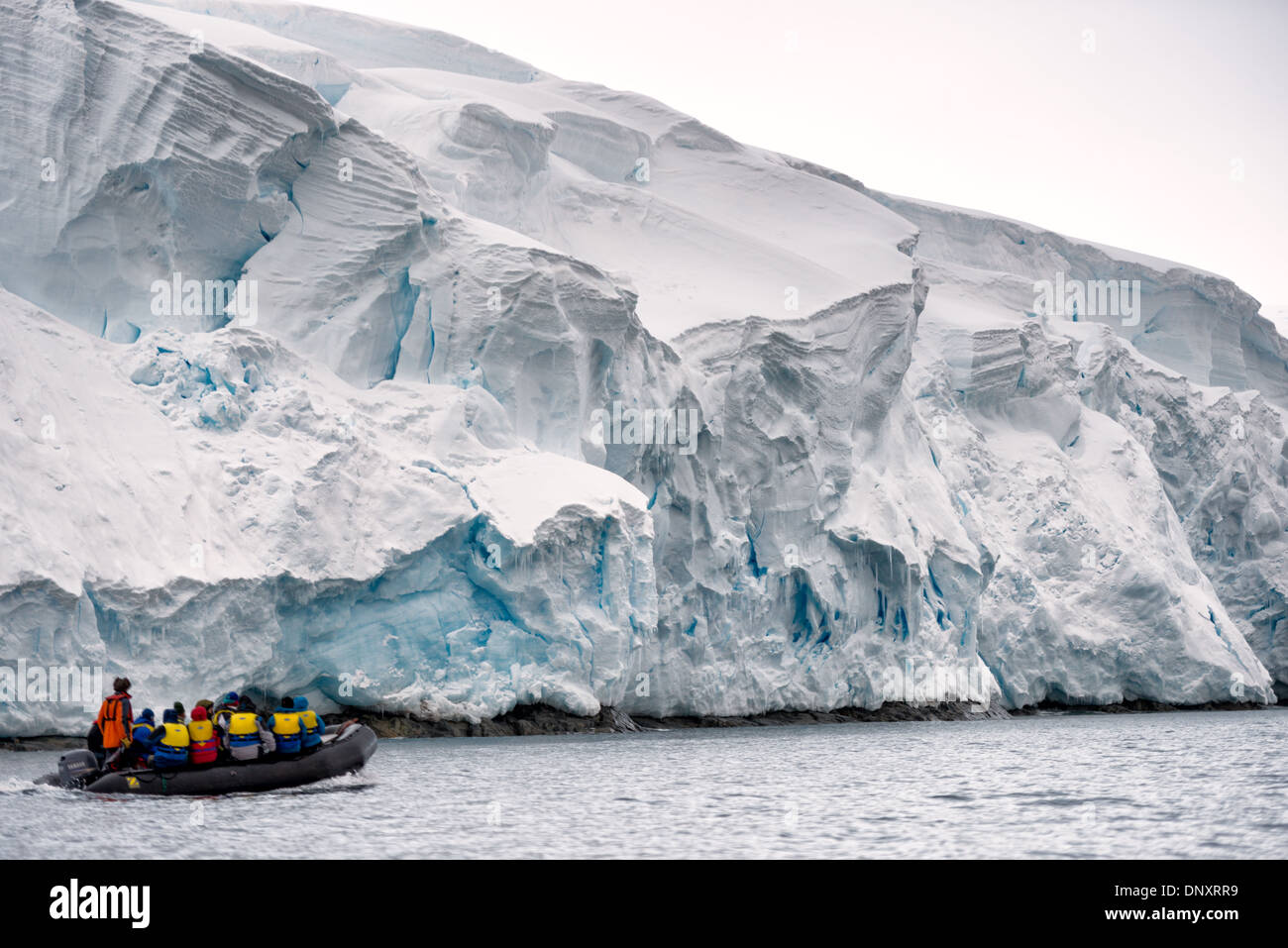Curtis Bay Glacier Cliffs Ice Boat Zodiac Peninsula Antarctic with People / CURTIS BAY, Antarctique — les écotouristes dans un bateau pneumatique Zodiac passent sous une falaise de glace glaciaire à Curtis Bay sur la péninsule Antarctique. La couleur bleue visible dans les crevasses et les zones de fracture révèle une glace si dense qu'elle absorbe les longueurs d'onde rouges et diffuse le bleu - un signe que le glacier a comprimé des siècles de chutes de neige en cristal sans pratiquement plus de bulles d'air. Banque D'Images