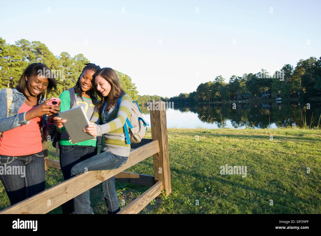 Pre-teen girls sitting on fence, à la tablette à l'ordinateur et téléphone cellulaire, à l'extérieur Banque D'Images