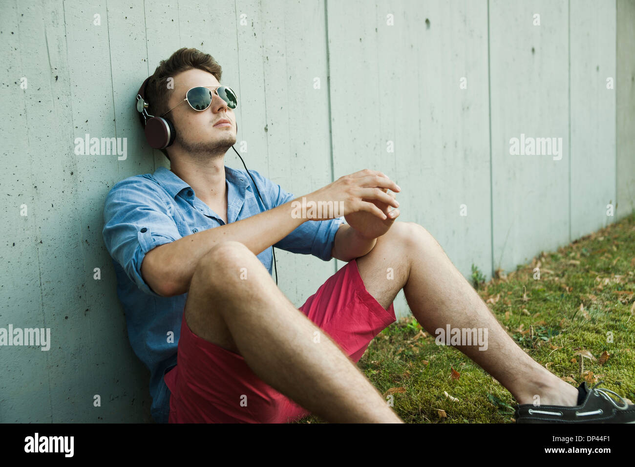 Jeune homme assis à côté de mur du bâtiment à l'extérieur, portant des écouteurs et des lunettes de soleil, Allemagne Banque D'Images