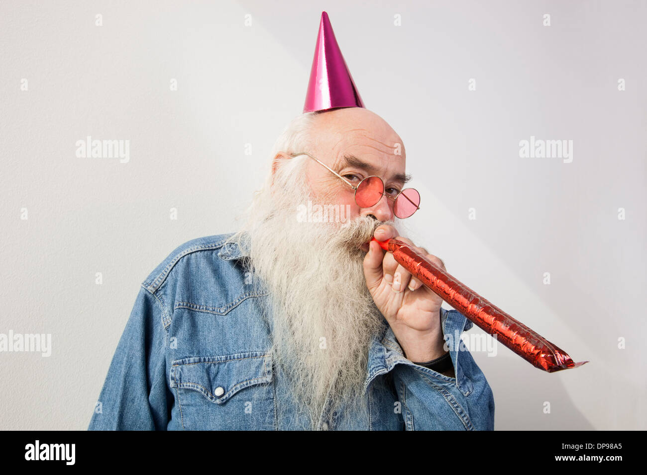 Portrait of senior man wearing party hat tout en soufflant corne contre fond gris Banque D'Images