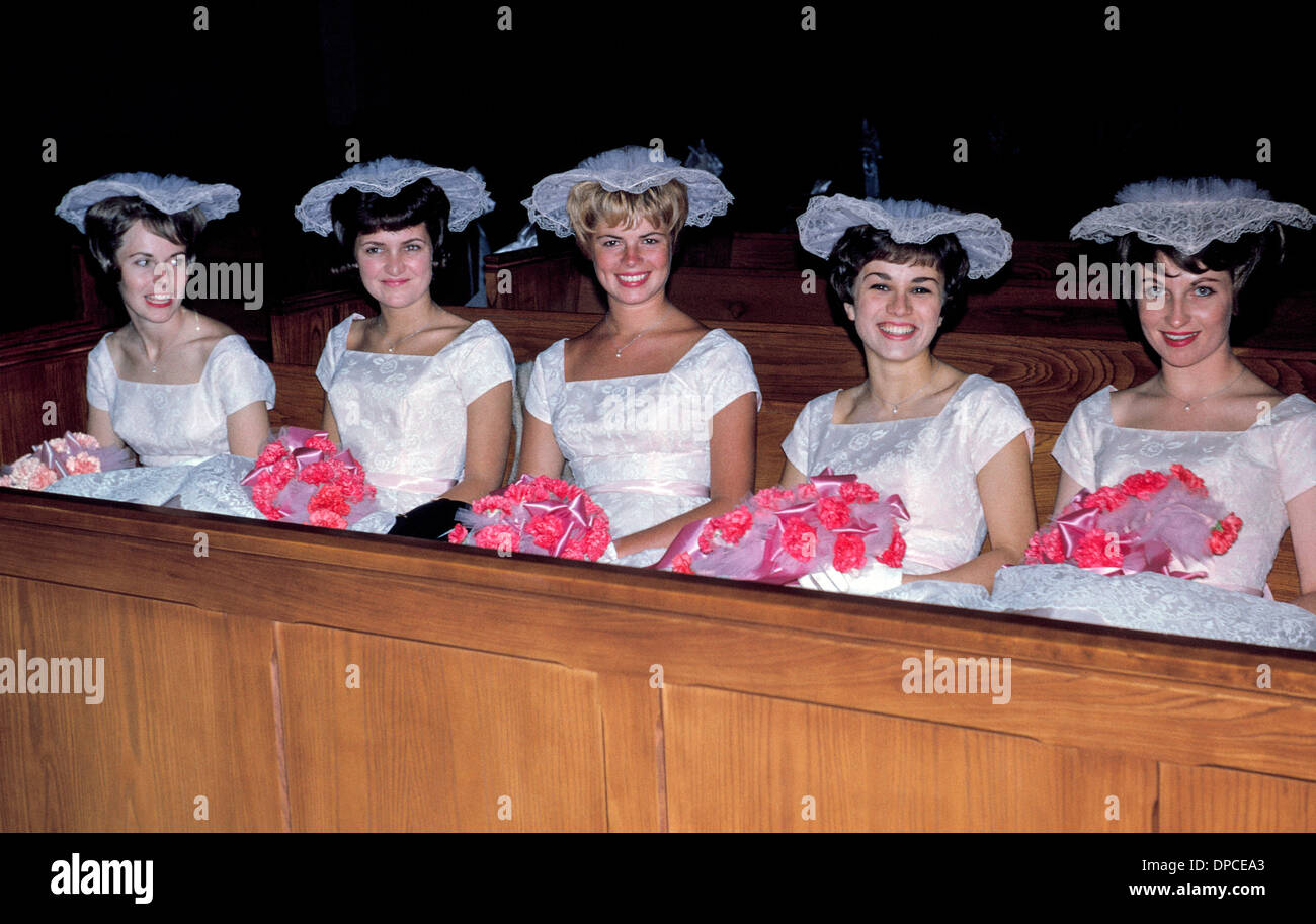 Cinq smiling demoiselles en robes blanches et des chapeaux à froufrous attendre dans une église d'un pew 1960 Cérémonie de mariage pour commencer. Banque D'Images