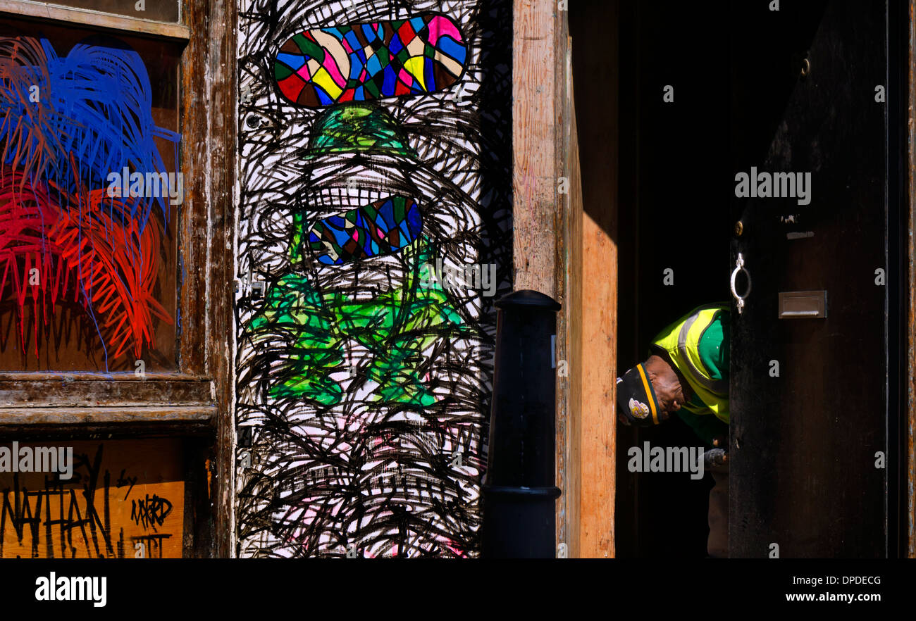 Un ouvrier de la réparation d'un cadre de porte à côté d'une fenêtre couverte de rue urbaine de cayenne en zone graffiti Brick Lane East London E1 UK Banque D'Images
