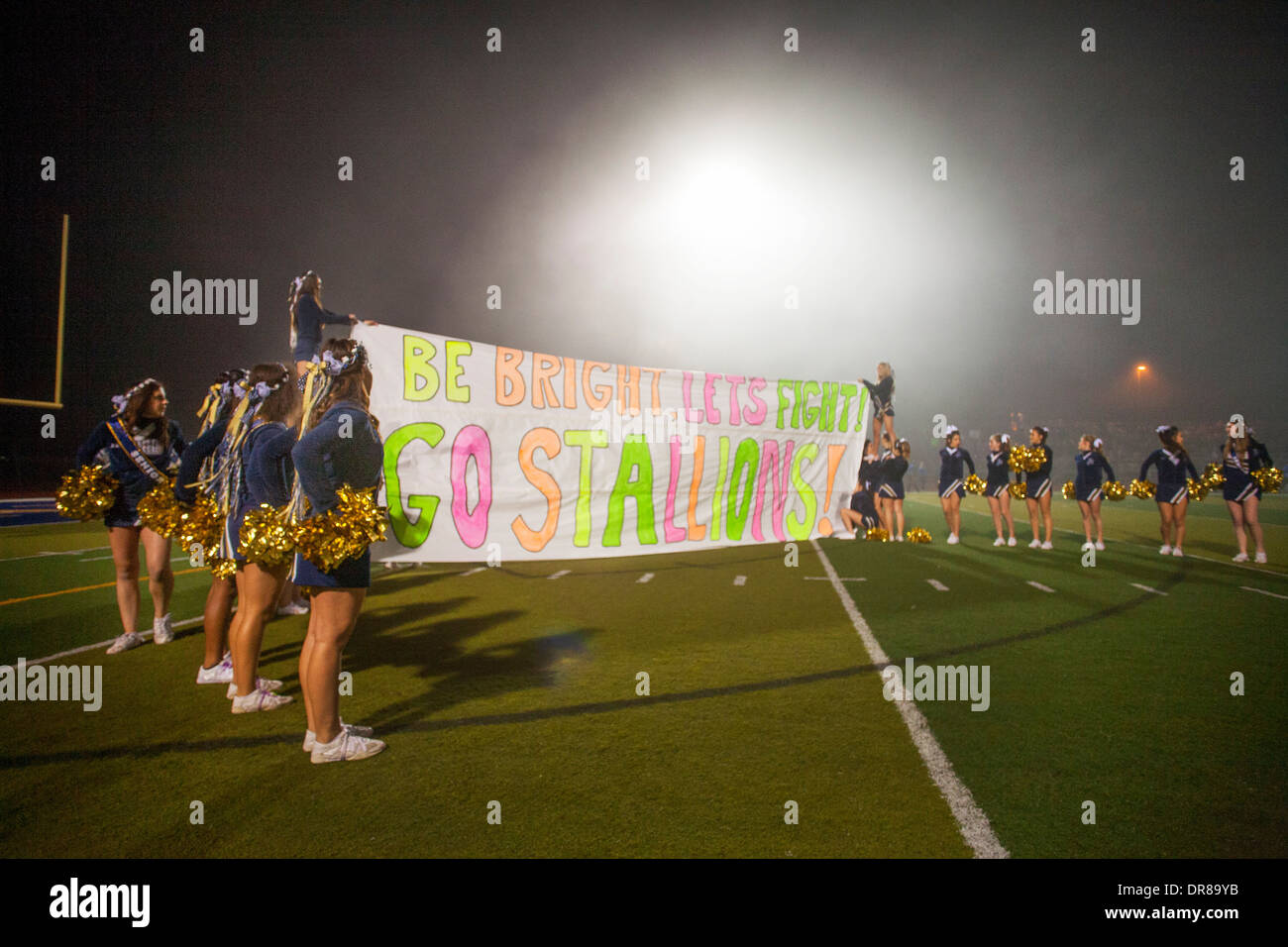 High school cheerleaders soulèvent une bannière d'inspiration pour leur équipe de football devant un jeu de nuit à San Juan Capistrano, CA. Banque D'Images