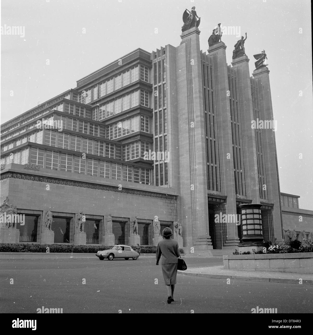 Années 1960, historique, une dame marchant jusqu'au Palais du Centenaire du Centre des expositions de Bruxelles (Palais des expositions), un grand bâtiment art déco à Bruxelles, en Belgique construit pour l'exposition universelle de 1935. Garé à l'extérieur d'une voiture Citroën. Banque D'Images