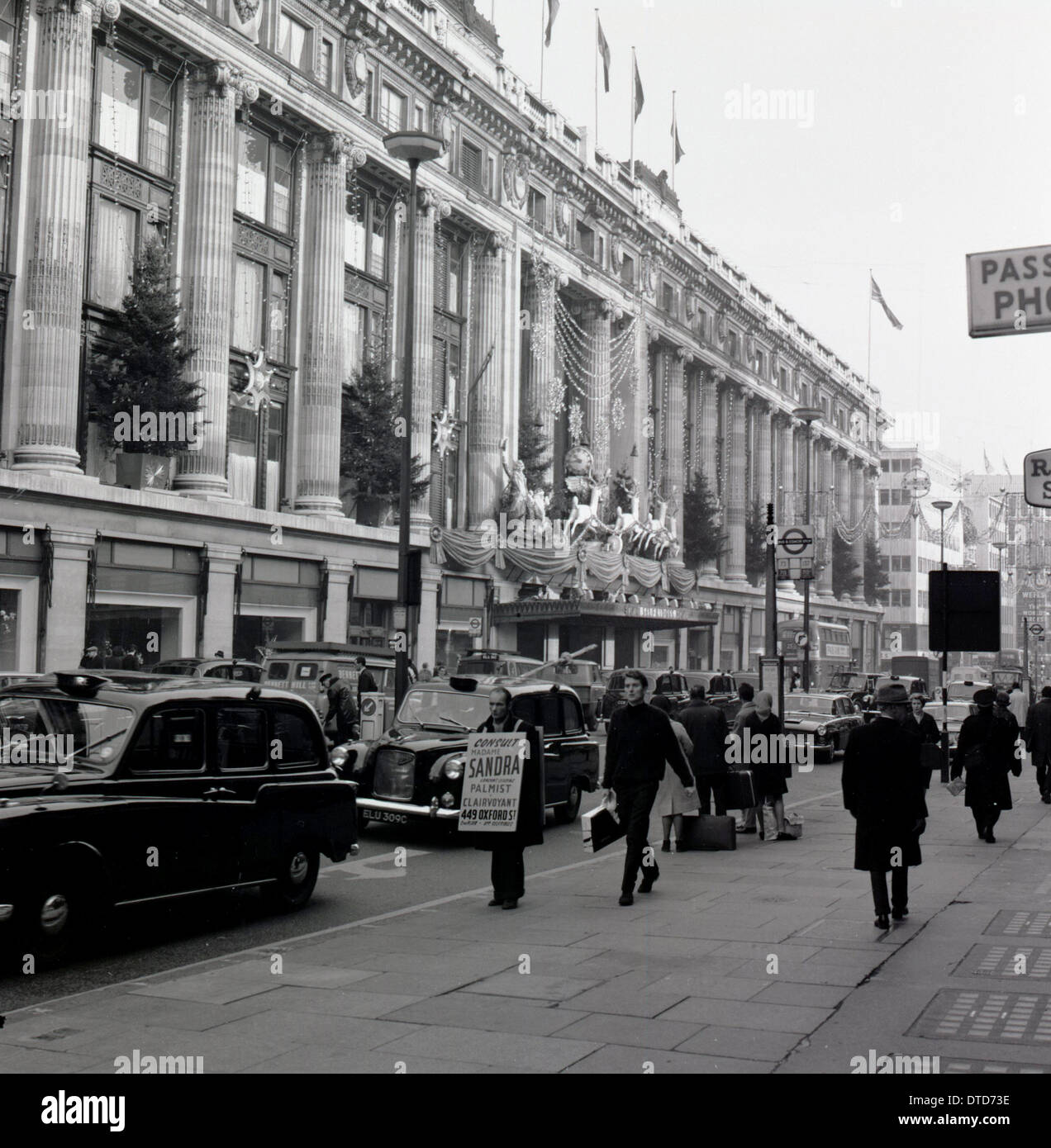 Années 1960, historique, décorations de Noël sur le grand magasin Selfridges sur une rue animée d'Oxford Street, Londres, Angleterre. Circulation très fréquentée avec taxis londoniens dans la rue. Sur le trottoir, un homme avec une planche sandwich. Banque D'Images