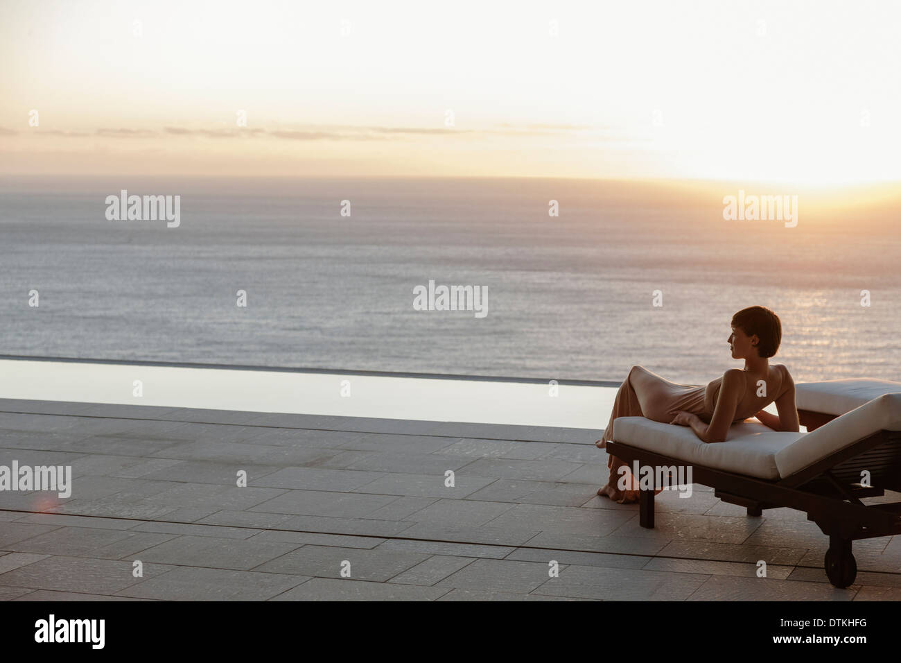 Woman in dress portant sur chaise de salon sur patio donnant sur l'océan au coucher du soleil Banque D'Images