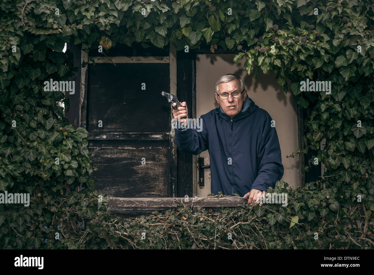 Furious senior man gesturing with a gun protéger sa propriété, debout devant des vieux envahi par la cabine. Banque D'Images
