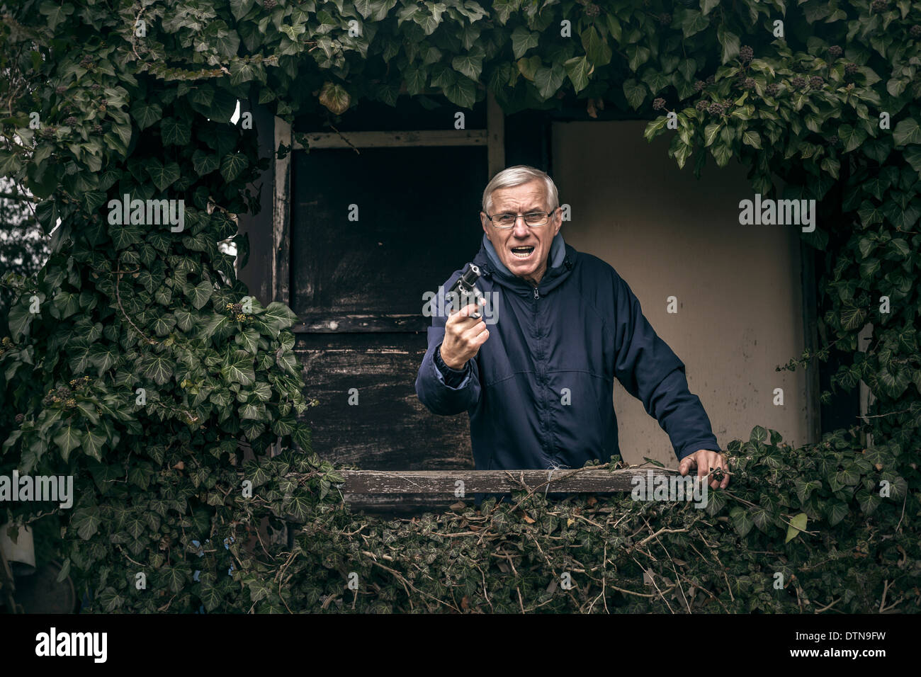 Furious senior man gesturing with a gun protéger sa propriété, debout devant des vieux envahi par la cabine. Banque D'Images