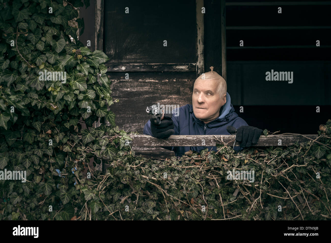 Senior homme masqué évadé visant une arme à feu, se cachant sur de vieux porche envahi par la cabine. Banque D'Images