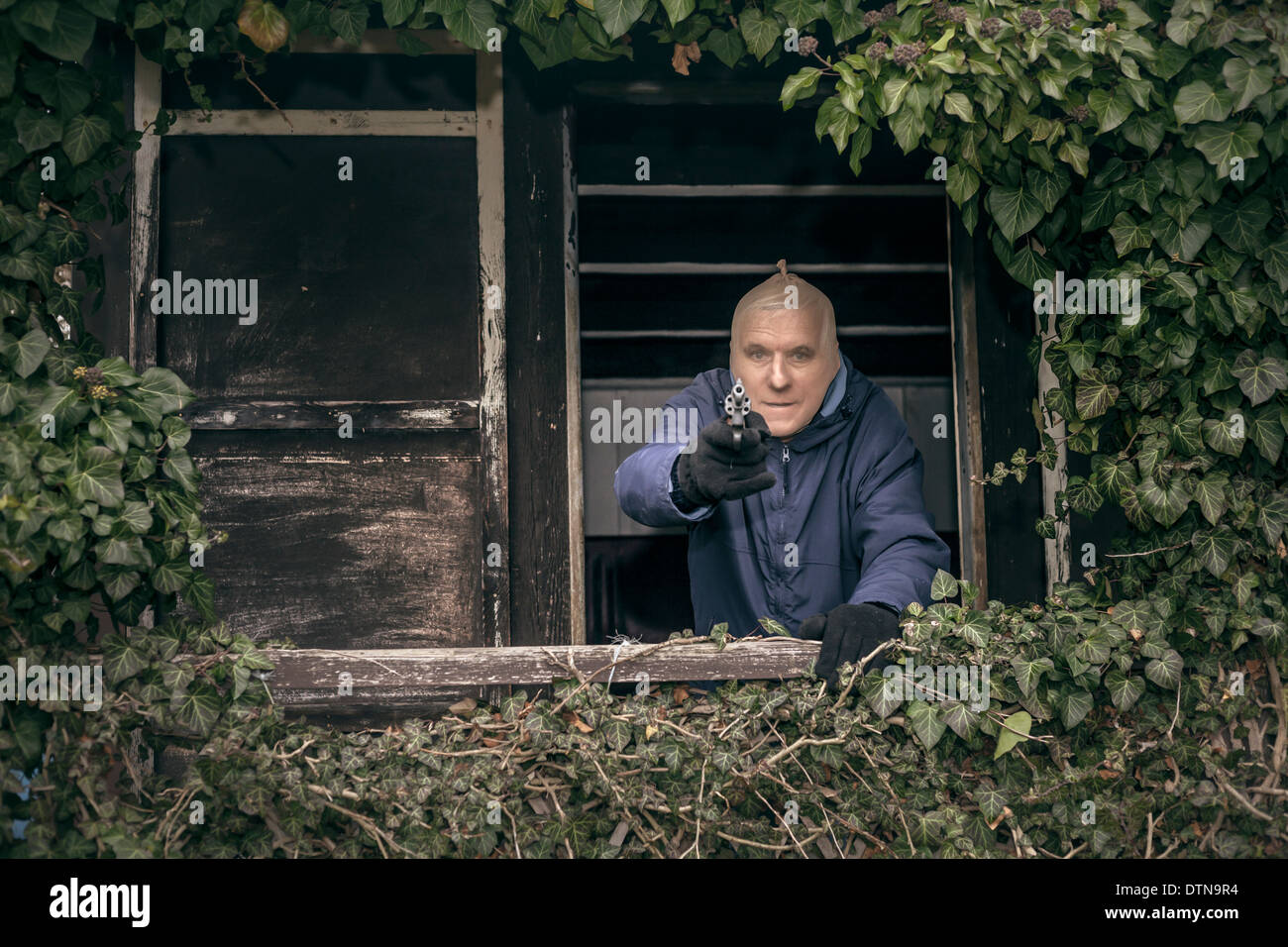 Senior homme masqué évadé visant une arme à feu, se cachant sur de vieux porche envahi par la cabine. Banque D'Images