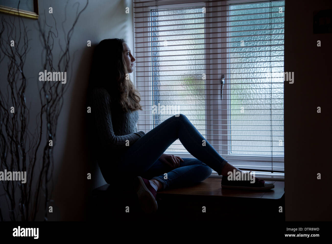 Jeune femme aux cheveux longs portant des jeans, triste et à la recherche d'une fenêtre, assis dans l'obscurité à la maison. Vue latérale. Banque D'Images