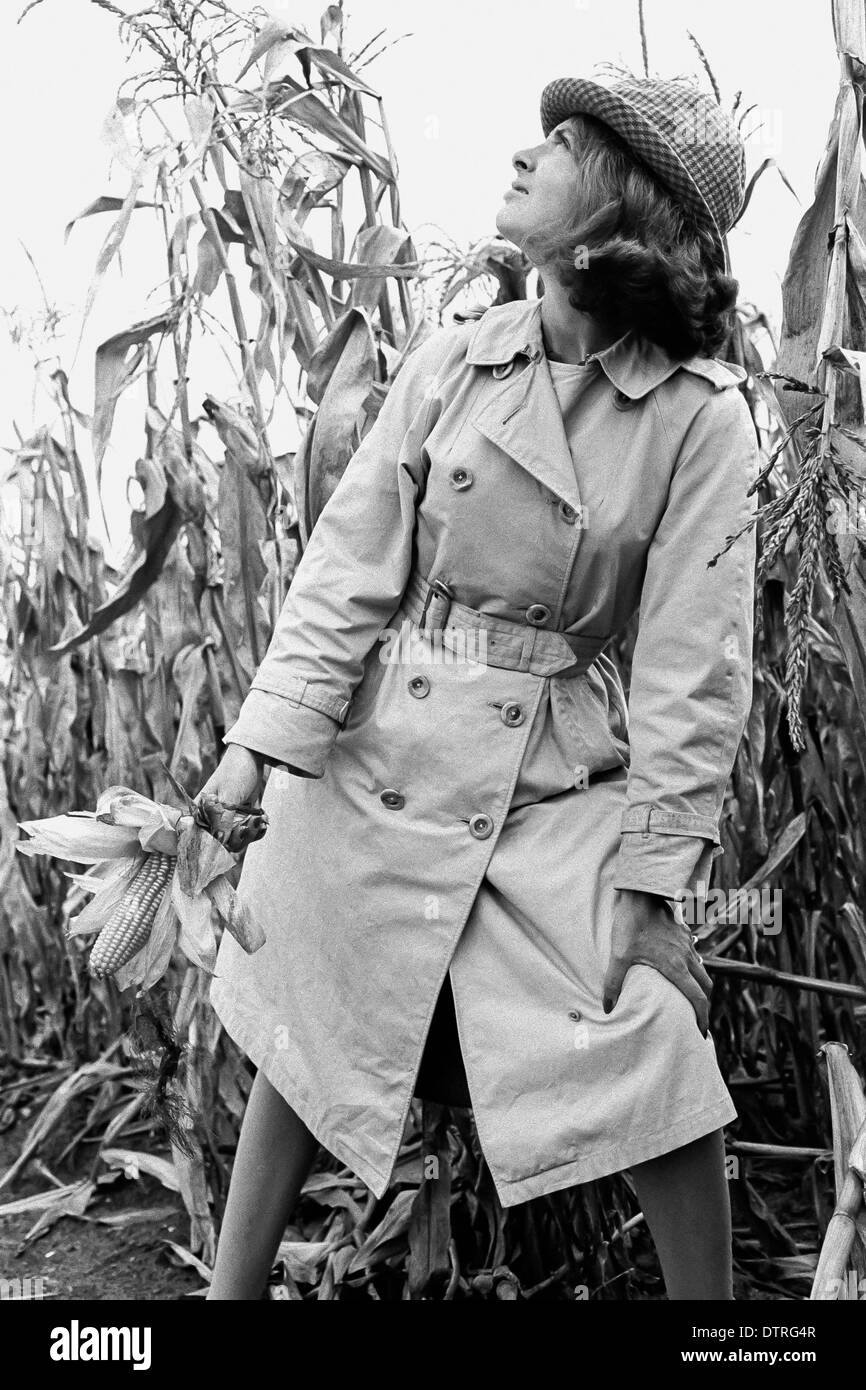 Modèle de mode des années 60 avec l'imperméable et le chapeau de poser dans un champ de maïs Banque D'Images