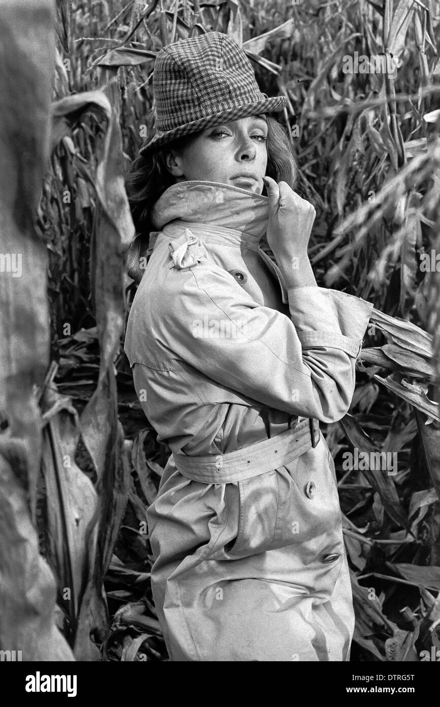 Modèle de mode des années 60 avec l'imperméable et le chapeau de poser dans un champ de maïs Banque D'Images