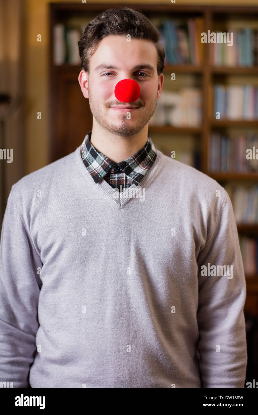 Beau jeune homme souriant avec nez de clown rouge, debout dans un salon Banque D'Images