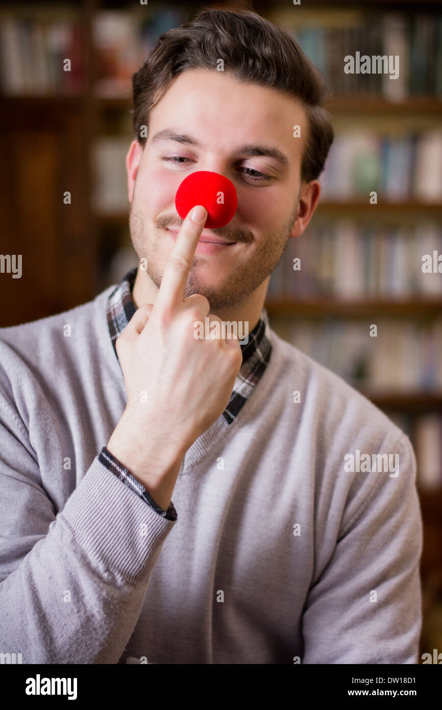 Beau jeune homme souriant et toucher le nez de clown rouge, debout dans un salon Banque D'Images