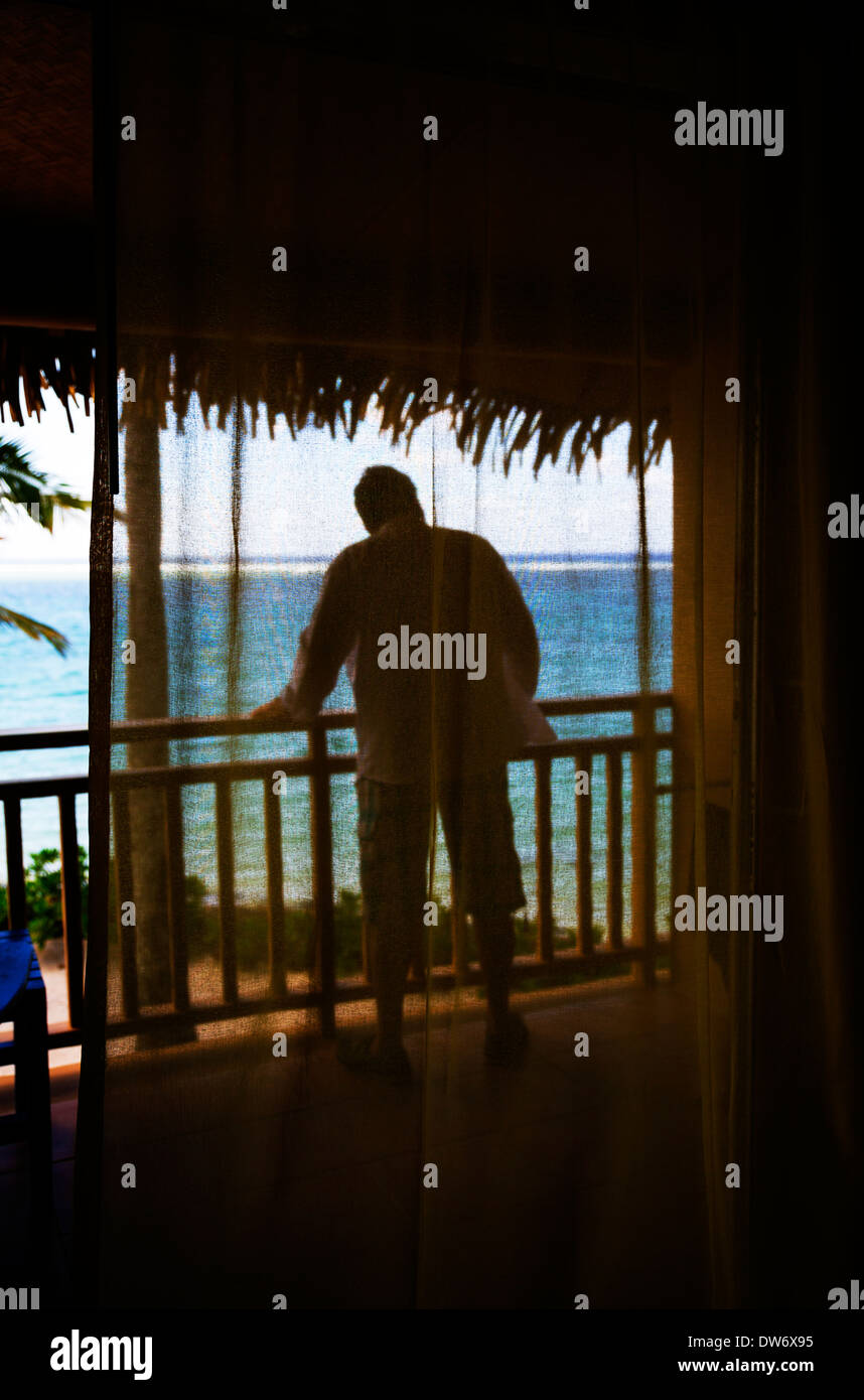 Un homme sur le site sur le balcon d'un hôtel. Silhouetted against a blue lagoon sur une île tropicale. Banque D'Images