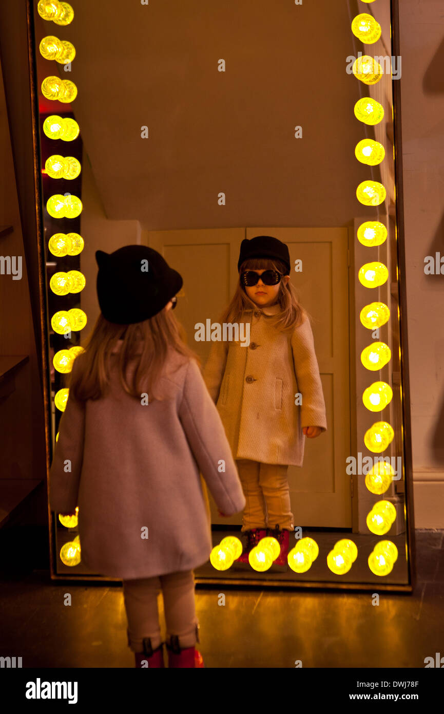 Une petite fille à lunettes à son reflet dans un miroir entouré par lighbulbs Banque D'Images