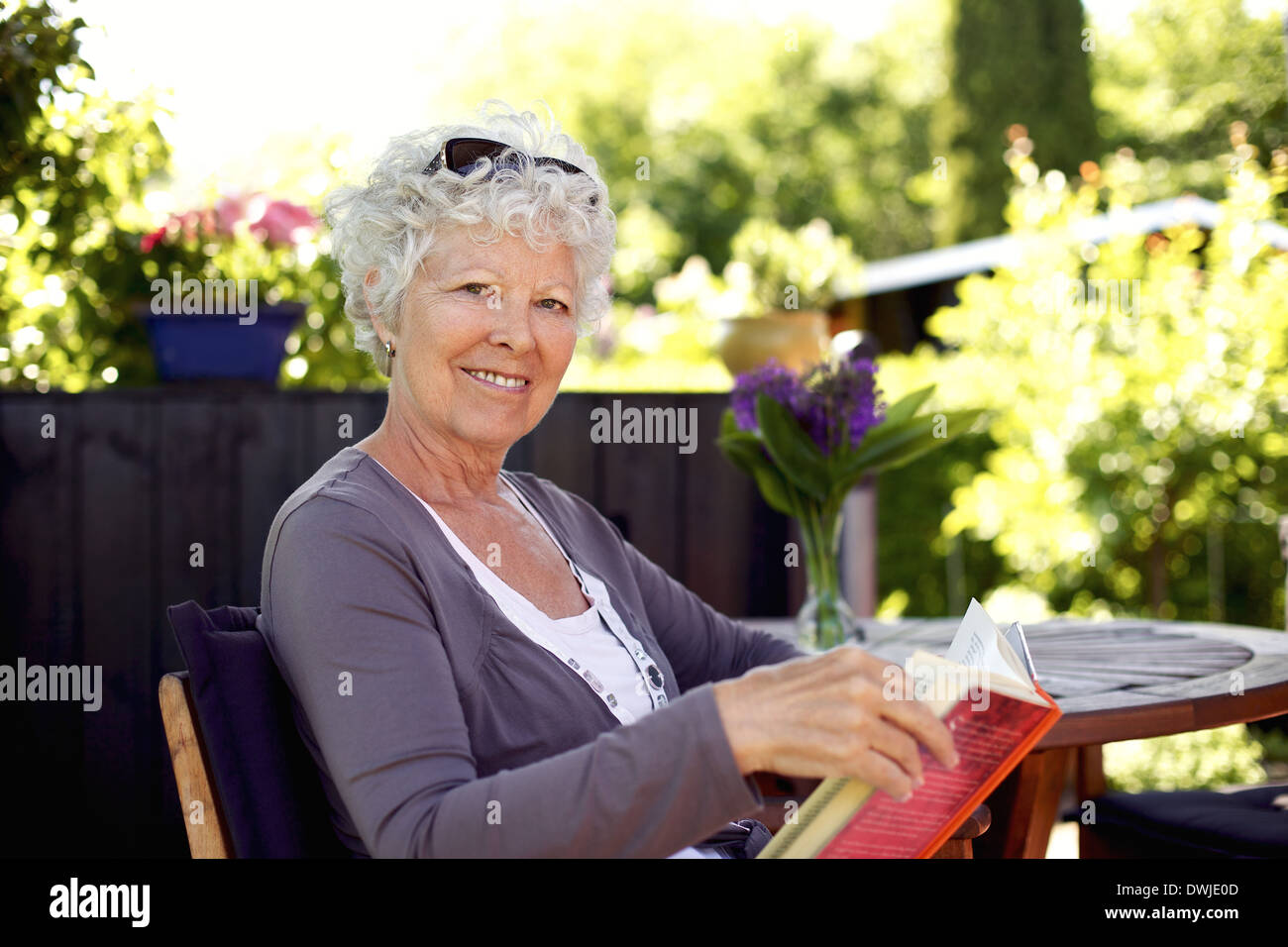 Hauts femme assise sur une chaise de jardin en tenant un livre à la caméra et au sourire Banque D'Images