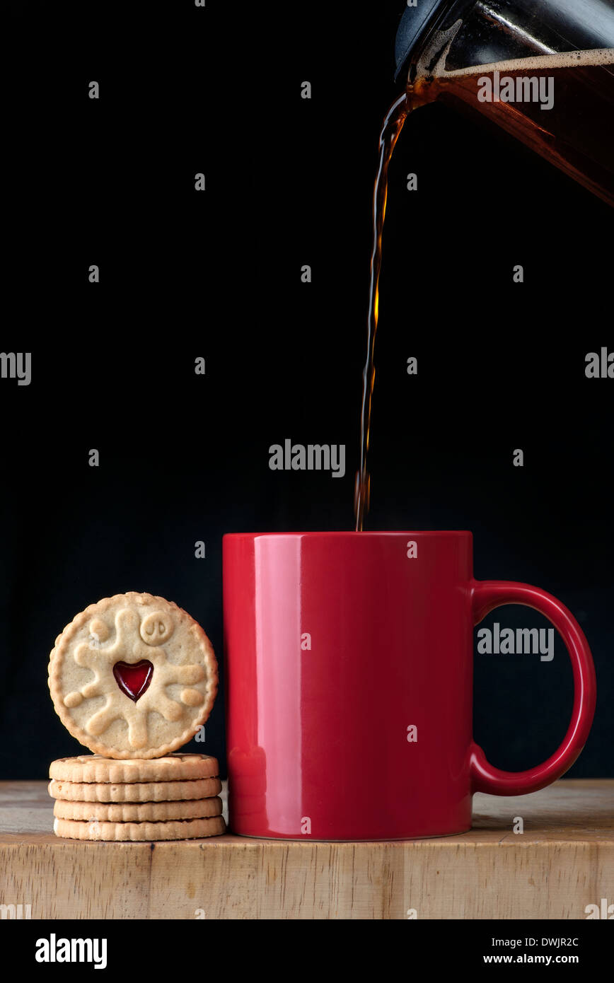 Verser du café d'une cafetière dans une tasse rouge et biscuits Banque D'Images